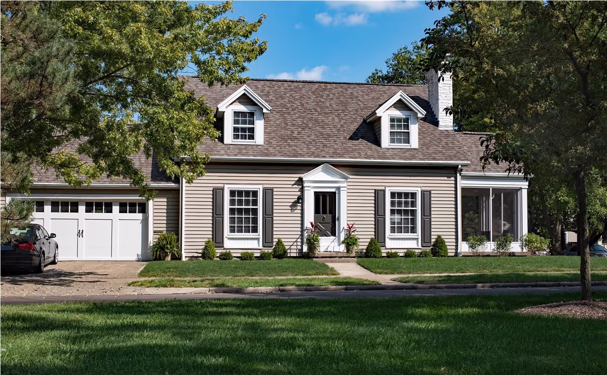 Beige Cape Cod-style house with dormers, black shutters and white windows behind a green lawn